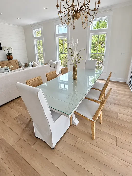 Elegant dining area with a glass table, white and wicker chairs, chandelier, and large windows with garden view.