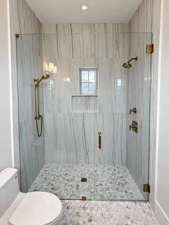 Modern bathroom with glass shower, brass fixtures, marble walls, hexagonal floor tiles, and a window.